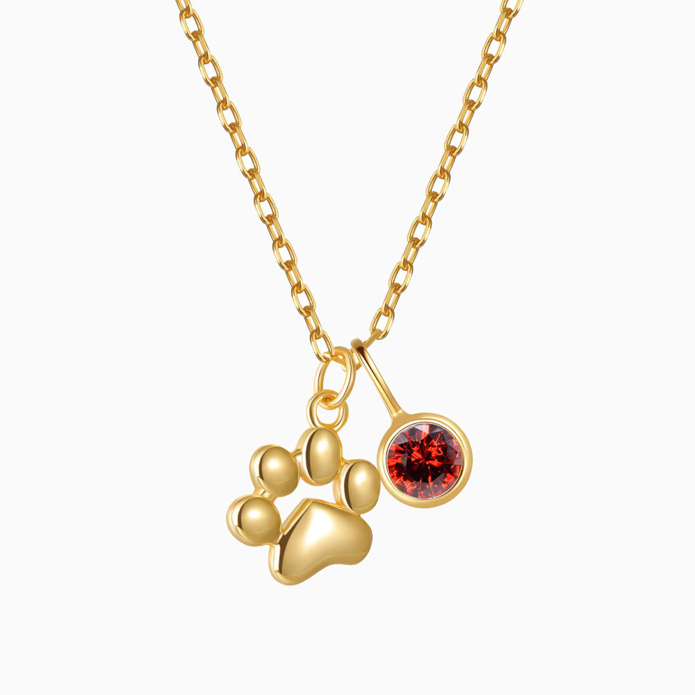 A gold-colored Teeny Tiny Paw Necklace with a red birthstone charm attached to a chain.